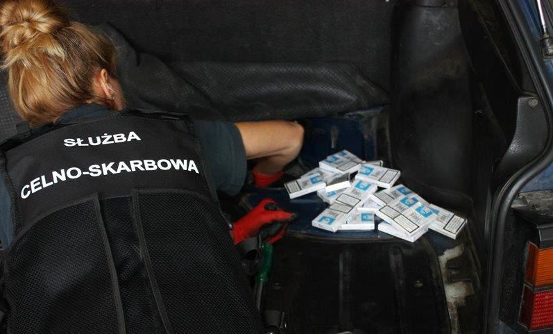 Funkcjonariuszka Służby Celno-Skarbowej przeszukuje bagażnik samochodu osobowego. Po jej prawej stronie leży sterta paczek papierosów.