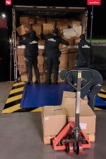 Dwóch funkcjonariuszy Służby Celno-Skarbowej pakuje kartony do naczepy samochodu ciężarowego. Trzeci zdejmuje karton z palety ustawionej na ręcznym wózku widłowym.