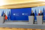 Przed mównicami stoją trzy osoby, kobieta po lewej, dwóch mężczyzn po prawej. Za nimi baner Ministerstwa Zdrowia i Ministerstwa Finansów oraz flagi Polski i Unii Europejskiej.