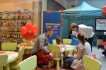 Zdjęcie przedstawia stanowisko w galerii handlowej w Olsztynie oraz stoliki z kolorowankami, zagadkami i mini gadżetami dla dzieci