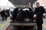 Funkcjonariusz KAS stoi przy przeszukiwanym samochodzie. W otwartym bagażniku widać rozkręcone elementy karoserii. Zatrzymany skuty kajdankami siedzi na stołku obok auta.